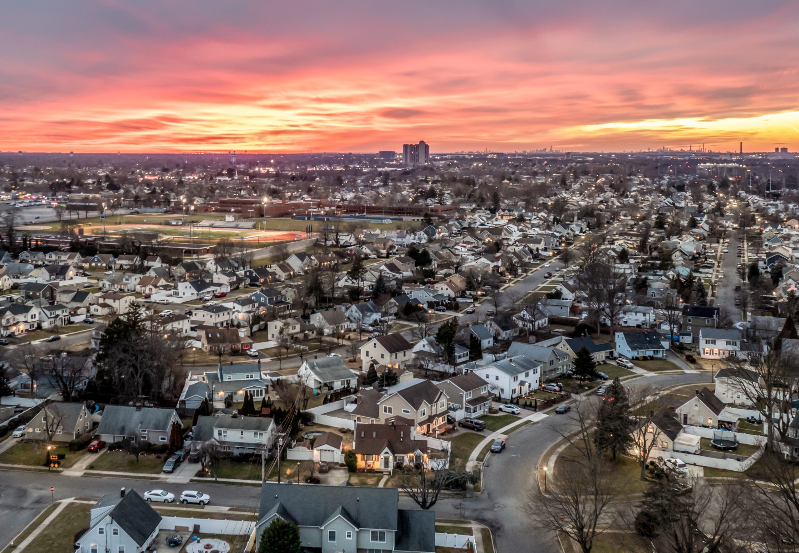 A sweeping aerial view of a suburban neighborhood in Nassau County, Long Island, captured at sunset. Rows of homes with driveways and tree-lined streets stretch across the frame, with a brightly lit sports field visible near the center. The mix of calm suburban streets and the vibrant sunset creates a striking view of everyday life in the NY Metro