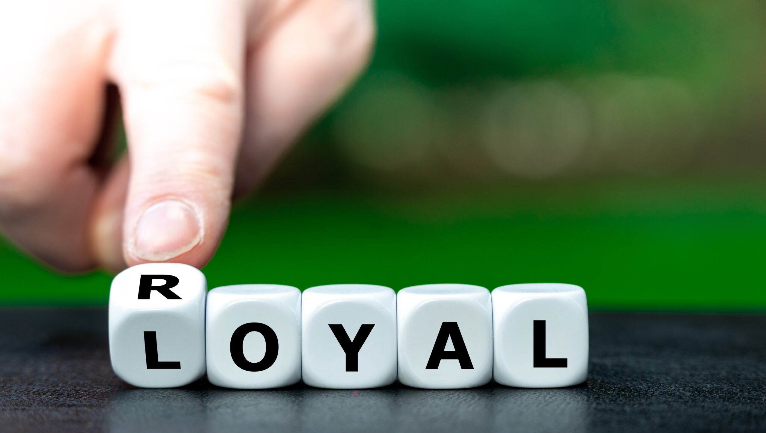 This image shows a close-up of white lettered dice spelling out the word 'LOYAL.' A hand is turning the first die, which shows the letter R, suggesting a shift between 'ROYAL' and 'LOYAL.' The background is blurred green, keeping the focus on the dice and the message of loyalty and royalty.